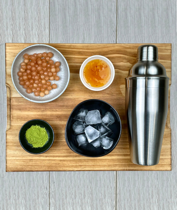 Silver, brushed stainless steel shaker displayed with ceramic dishes containing cooked Red Grapefruit pearls, Yuzu fruit mix, Matcha powder, ice