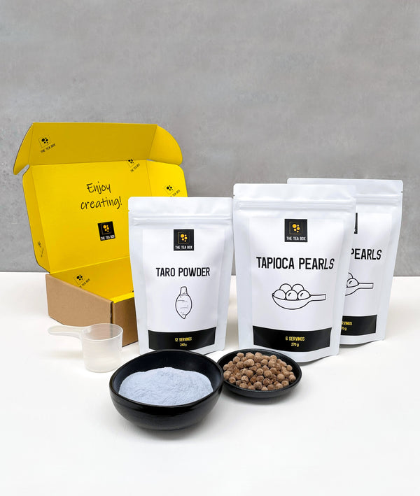 Contents of a 12 pack Taro Bubble Tea Kit. Taro powder and brown tapioca pearls in pouches, taro powder and uncooked brown tapioca pearls displayed in ceramic dishes, scoop, cardboard gift box