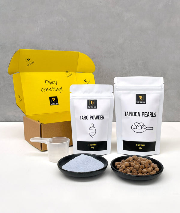 Contents of a 3 pack Taro Bubble Tea Kit. Taro powder and brown tapioca pearls in pouches, taro powder and uncooked brown tapioca pearls displayed in ceramic dishes, scoop, cardboard gift box