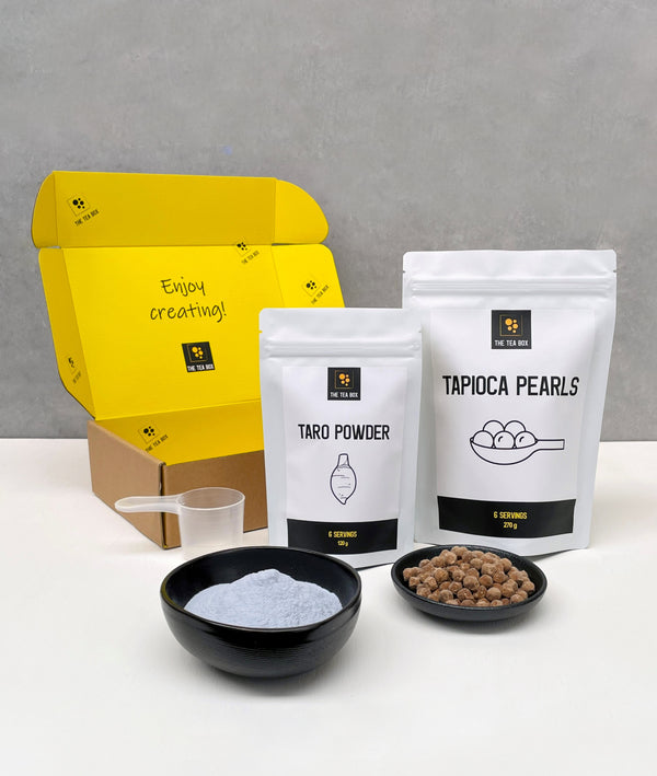 Contents of a 6 pack Taro Bubble Tea Kit. Taro powder and brown tapioca pearls in pouches, taro powder and uncooked brown tapioca pearls displayed in ceramic dishes, scoop, cardboard gift box