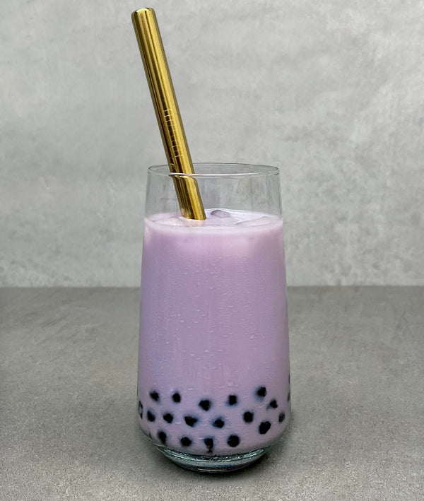 A glass of vibrant purple Taro Bubble Tea with Brown Tapioca Pearls, ice cubes and a gold stainless steel bubble tea straw